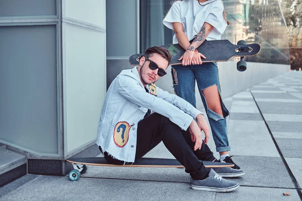 Şık giyimli çift - bir longboard ve kız arkadaşı açık havada duran üzerinde oturan genç hipster adam. — Stok fotoğraf