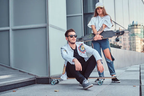 Módní oblečený pár - mladý bokovky muž sedí na longboard a jeho přítelkyně, stojící poblíž venku. — Stock fotografie