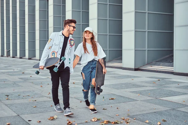 Feliz pareja joven cogidos de la mano y caminando juntos con patinetas en la calle moderna en tiempo ventoso . — Foto de Stock