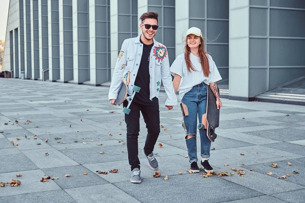 Feliz pareja joven cogidos de la mano y caminando juntos con patinetas en la calle moderna en tiempo ventoso . —  Fotos de Stock