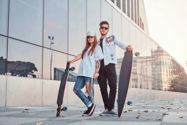 Atraente jovem casal vestido com roupas da moda posando com skates perto do arranha-céu . — Fotografia de Stock