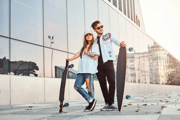 Aantrekkelijke jonge paar gekleed in trendy kleding poseren met skateboards in de buurt van wolkenkrabber. — Stockfoto