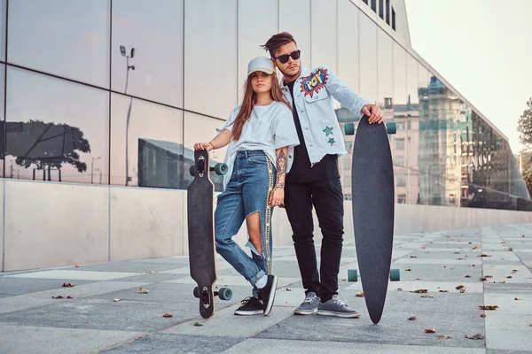 Приваблива молода пара, одягнена в модний одяг, позує зі скейтбордами біля хмарочоса . — стокове фото