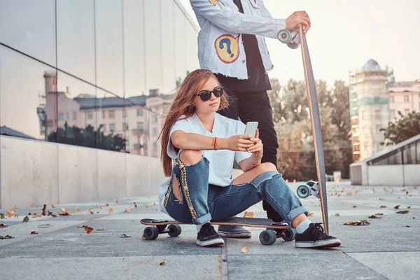 Joven chica hipster sentado en longboard y el uso de un teléfono inteligente mientras su novio de pie cerca . — Foto de Stock