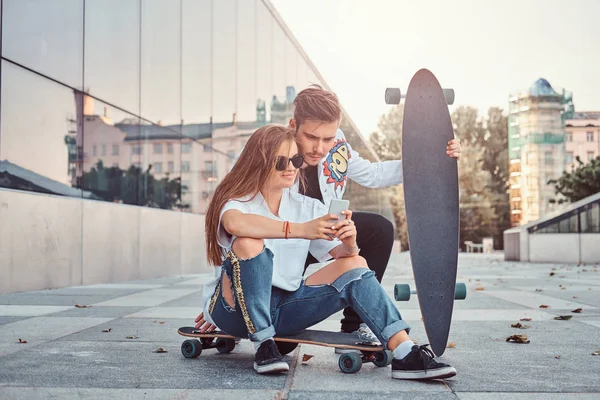 Pareja vestida de moda con skateboards ve fotos interesantes juntos en el teléfono inteligente en la calle . — Foto de Stock