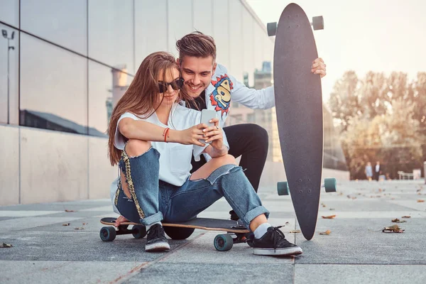 Pareja vestida de moda con skateboards ve fotos interesantes juntos en el teléfono inteligente en la calle . — Foto de Stock
