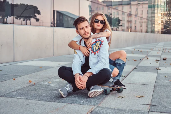 Trendy gekleidetes junges Paar - hübsches Mädchen umarmt ihren Freund, während sie zusammen auf einem Skateboard in der Nähe eines Wolkenkratzers sitzen. — Stockfoto