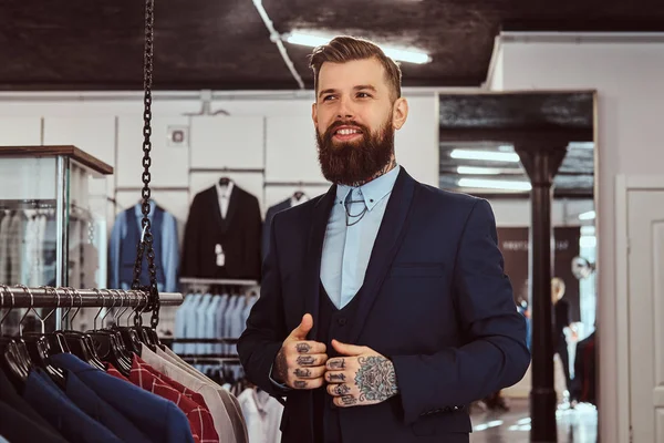 Maschio sorridente tatuato con barba elegante e capelli vestiti in abito elegante in piedi nel negozio di abbigliamento maschile . — Foto Stock