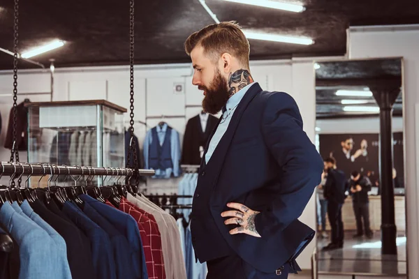 Elegantemente vestido barbudo macho com tatuagens nas mãos e pescoço escolhe novo traje na loja de moda masculina . — Fotografia de Stock