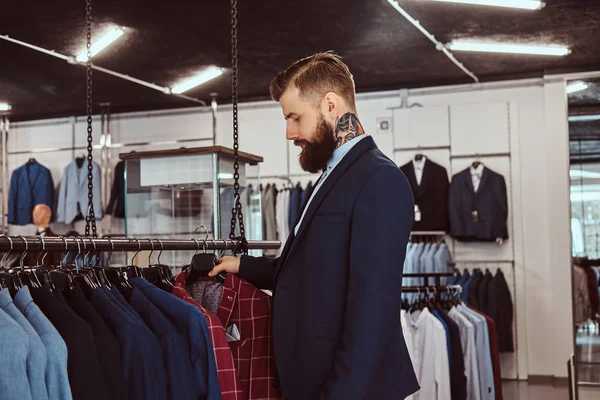 Elegantemente vestido barbudo macho com tatuagens nas mãos e pescoço escolhe novo traje na loja de moda masculina . — Fotografia de Stock