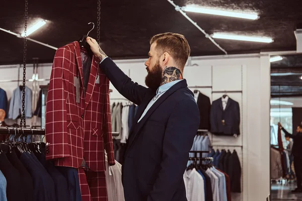 Elegancko ubrany Brodaty mężczyzna z tatuaży na rękach i szyi wybiera nowy kostium w magazynie odzieży męskiej. — Zdjęcie stockowe