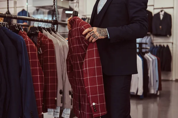 Foto ritagliata di un maschio elegante con il tatuaggio sulla mano tiene la nuova giacca in un negozio di abbigliamento maschile . — Foto Stock