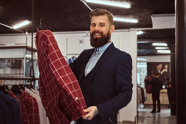 Elegantemente vestido barbudo macho com tatuagens nas mãos e pescoço escolhe novo traje na loja de moda masculina . — Fotografia de Stock