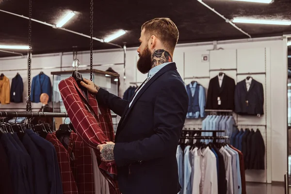 Elegantemente vestido barbudo macho com tatuagens nas mãos e pescoço escolhe novo traje na loja de moda masculina . — Fotografia de Stock