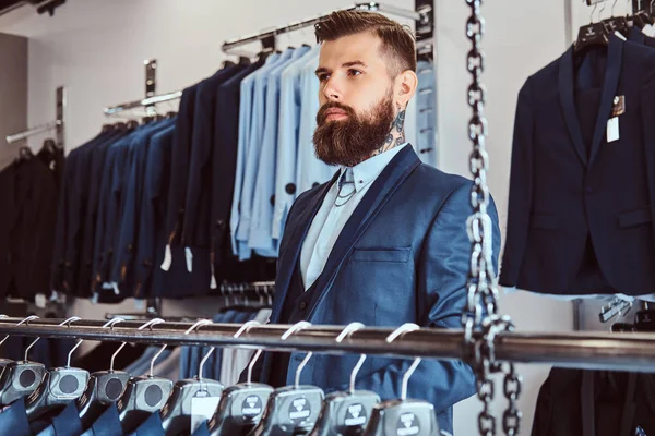 Homme tatoué pensif avec une barbe élégante et des cheveux habillés en costume élégant debout dans un magasin de vêtements pour hommes . — Photo