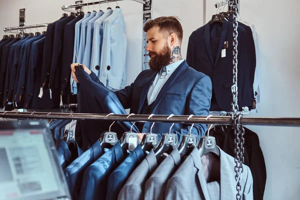 Elegantemente vestido barbudo macho com tatuagens nas mãos e pescoço escolhe novo traje na loja de moda masculina . — Fotografia de Stock