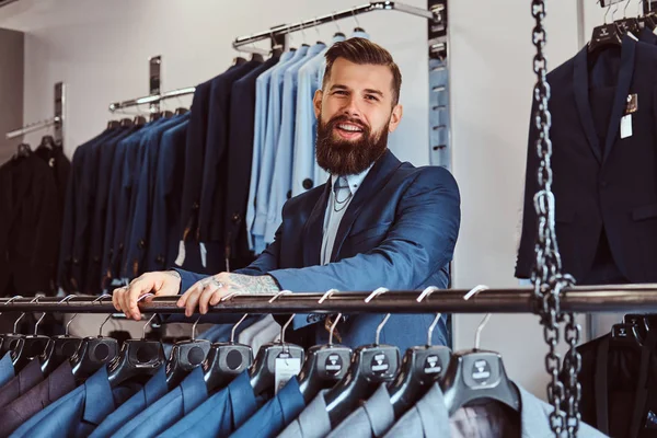 Sorrindo tatuado macho com barba elegante e cabelo vestido com terno elegante em pé em uma loja de moda masculina . — Fotografia de Stock