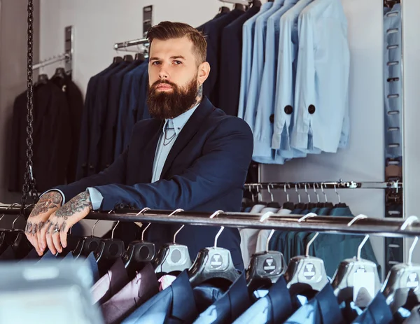 Maschio tatuato pensoso con barba elegante e capelli vestiti in abito elegante in piedi in un negozio di abbigliamento maschile . — Foto Stock