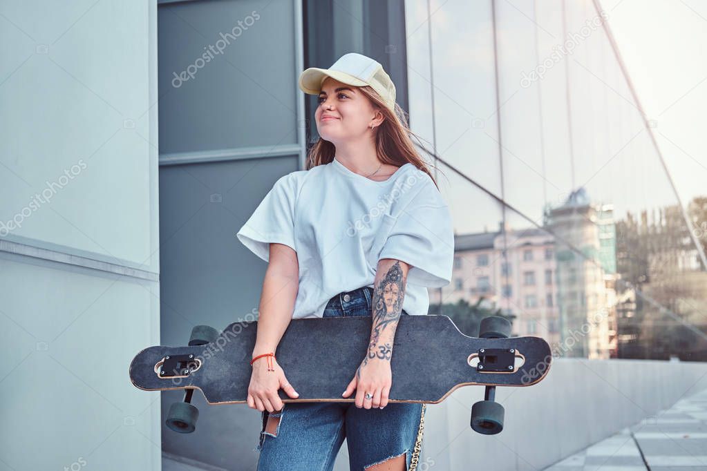 Young hipster girl in cap dressed in a white shirt and ripped jeans holds skateboard while posing near skyscraper.