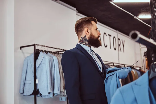 Elegantemente vestito maschio barbuto con tatuaggi su mani e collo sceglie il nuovo costume nel negozio di abbigliamento maschile . — Foto Stock