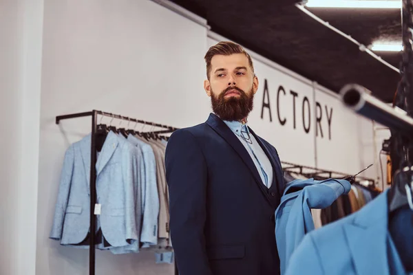 Elegant gekleed bebaarde man met tatoeages op handen en nek kiest nieuw kostuum in de herenkleding winkel. — Stockfoto