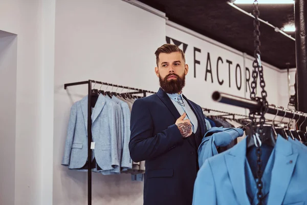 Elegantemente vestido barbudo macho com tatuagens nas mãos e pescoço escolhe novo traje na loja de moda masculina . — Fotografia de Stock