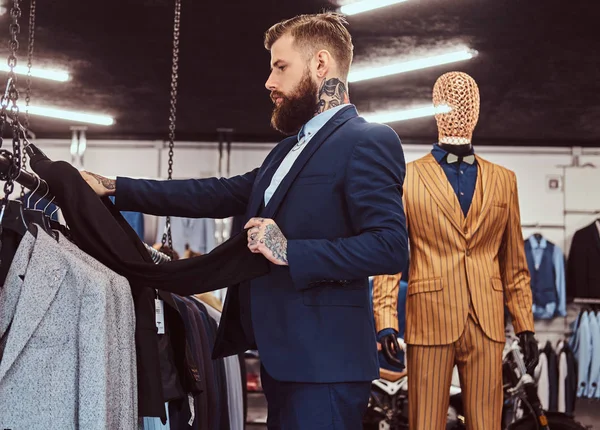 Elegantemente vestido barbudo macho com tatuagens nas mãos e pescoço escolhe novo terno em uma loja de moda masculina . — Fotografia de Stock