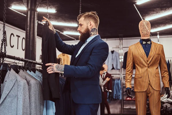 Elegantemente vestito maschio barbuto con tatuaggi su mani e collo sceglie nuovo abito in un negozio di abbigliamento maschile . — Foto Stock