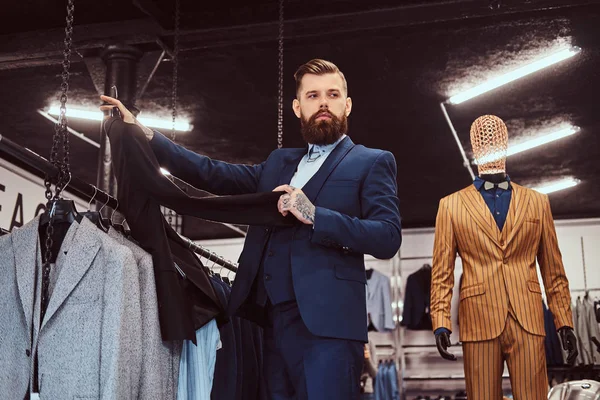 Elegantemente vestito maschio barbuto con tatuaggi su mani e collo sceglie nuovo abito in un negozio di abbigliamento maschile . — Foto Stock