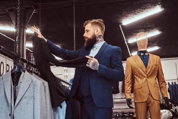 Elegant gekleideter bärtiger Mann mit Tätowierungen an Händen und Hals wählt neuen Anzug in einem Herrengeschäft. — Stockfoto
