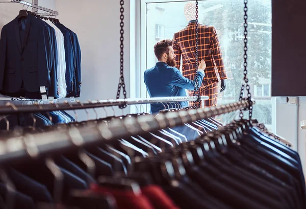 O vendedor barbudo elegante preocupa-se com o terno em um manequim em uma loja de roupa masculina . — Fotografia de Stock