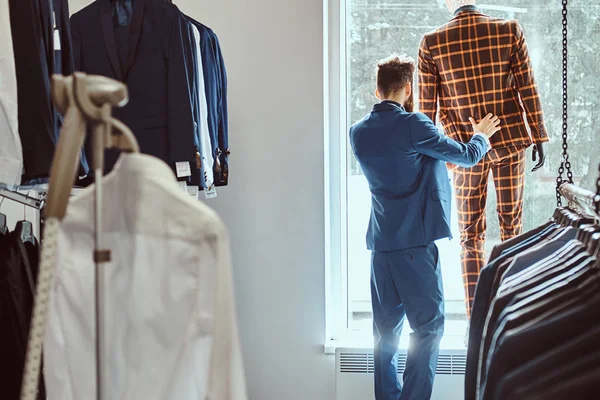 Elegante barbuto venditore cura di tuta su un manichino in un negozio di abbigliamento maschile . — Foto Stock