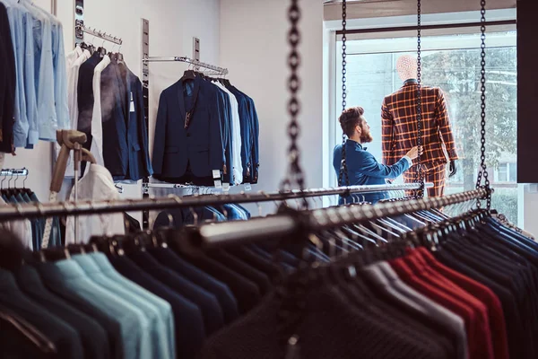 O vendedor barbudo elegante preocupa-se com o terno em um manequim em uma loja de roupa masculina . — Fotografia de Stock