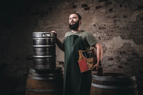Cervecero barbudo en delantal sostiene la caja de madera con cerveza mientras está cerca de barriles en la fábrica de cerveza . — Foto de Stock