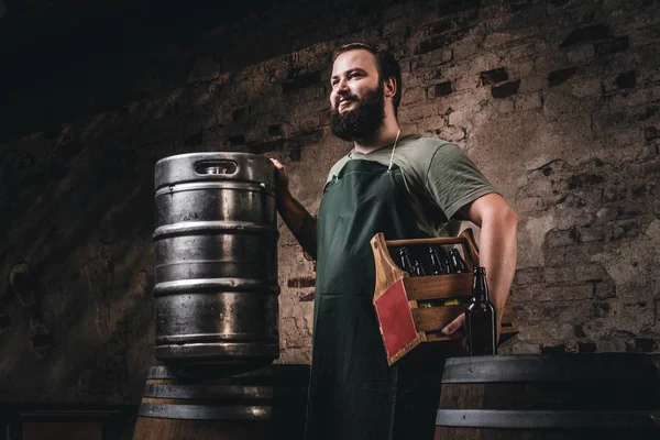 Cervecero barbudo en delantal sostiene la caja de madera con cerveza mientras está cerca de barriles en la fábrica de cerveza . — Foto de Stock