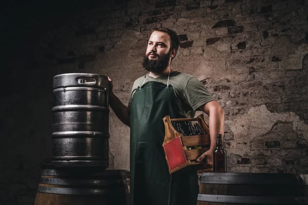 Bärtiger Brauer in Schürze hält die Holzkiste mit Bier in der Nähe von Fässern in der Brauereifabrik. — Stockfoto