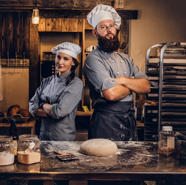 Chef with his assistant in cook uniform posing with crossed arms near table with ready dough in the bakery.