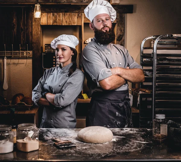 Шеф-повар со своим помощником в форме повара позирует со скрещенными руками возле стола с готовым тестом в пекарне . — стоковое фото
