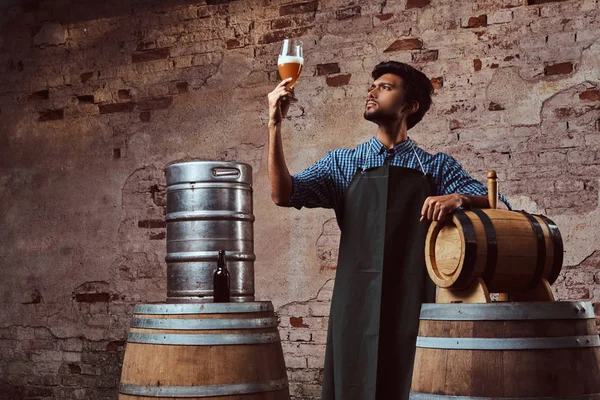 Cervecero experto en delantal de pie cerca de barriles que comprueban la calidad de la bebida elaborada en la fábrica de cerveza . — Foto de Stock