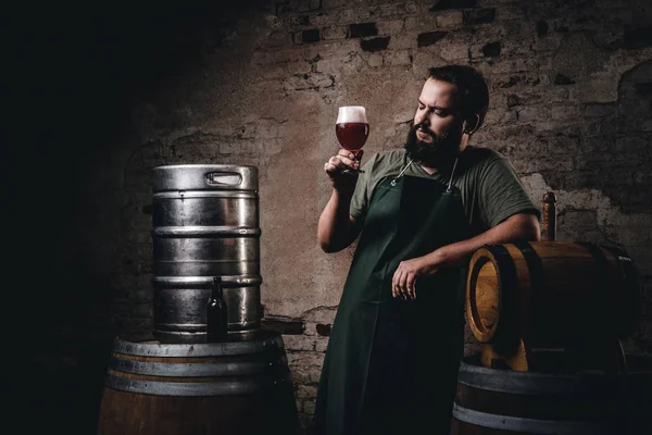 Cervecero en delantal de pie cerca de barriles y bebidas cerveza artesanal en la antigua fábrica de cerveza . — Foto de Stock