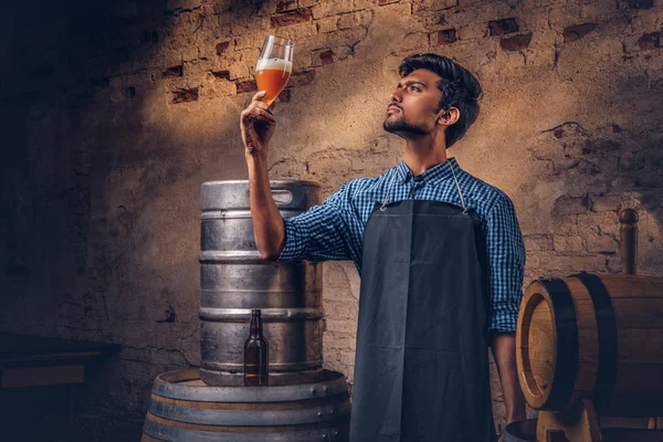 Cervecero experto en delantal de pie cerca de barriles que comprueban la calidad de la bebida elaborada en la fábrica de cerveza . — Foto de Stock