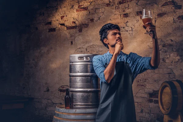 Cervecero experto en delantal de pie cerca de barriles que comprueban la calidad de la bebida elaborada en la fábrica de cerveza . — Foto de Stock