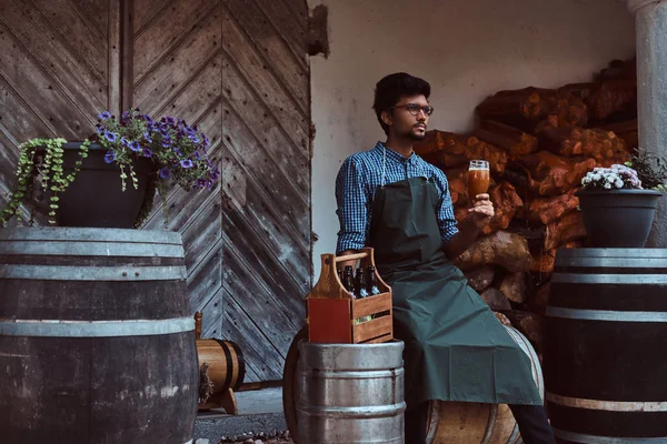 Brewmaster sitting on a wooden barrel and holds a glass of craft beer, relaxes after work.