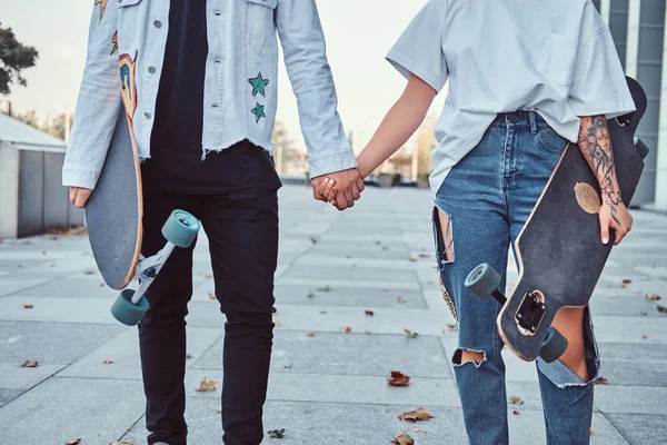 Foto recortada de jóvenes patinadores vestidos de moda tomados de la mano mientras están de pie cerca de rascacielos . —  Fotos de Stock