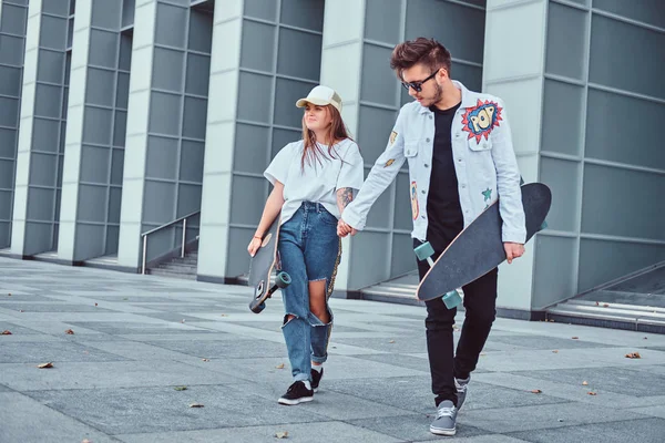 Gelukkige jonge paar houden handen en lopen samen met skateboards op moderne straat in winderig weer. — Stockfoto