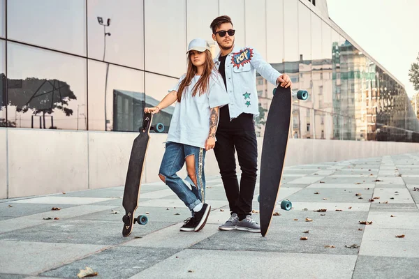 Attraktiva ungt par klädda i trendiga kläder poserar med skateboards nära skyskrapa. — Stockfoto
