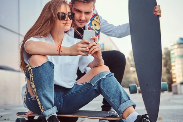 Pareja vestida de moda con skateboards ve fotos interesantes juntos en el teléfono inteligente en la calle . — Foto de Stock