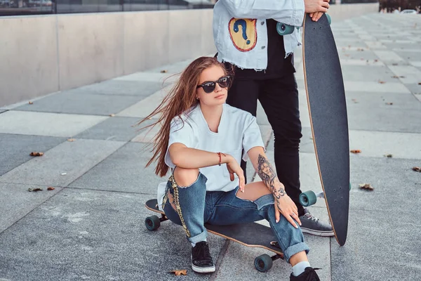 Hipster ragazza seduta su longboard mentre il suo ragazzo in piedi vicino all'aperto . — Foto Stock