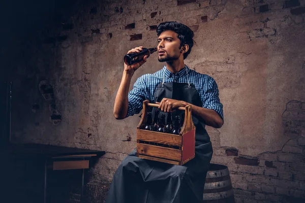 Brewmaster sentado en un barril de madera y sostiene un vaso de cerveza artesanal, se relaja después del trabajo . — Foto de Stock