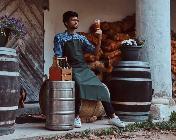 Brewmaster sentado en un barril de madera y sostiene un vaso de cerveza artesanal, se relaja después del trabajo . — Foto de Stock